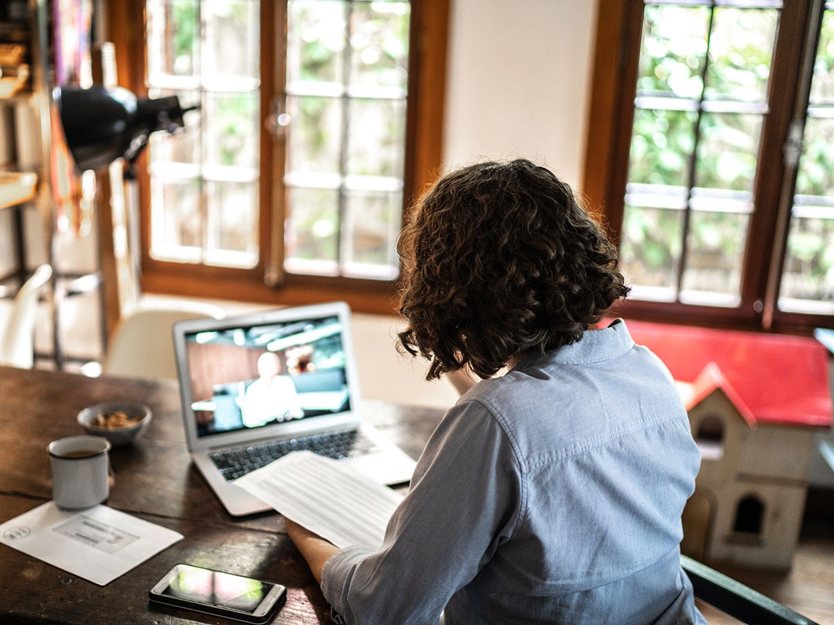 A woman with dark curly hair is sitting at a desk next to a window looking at her laptop.