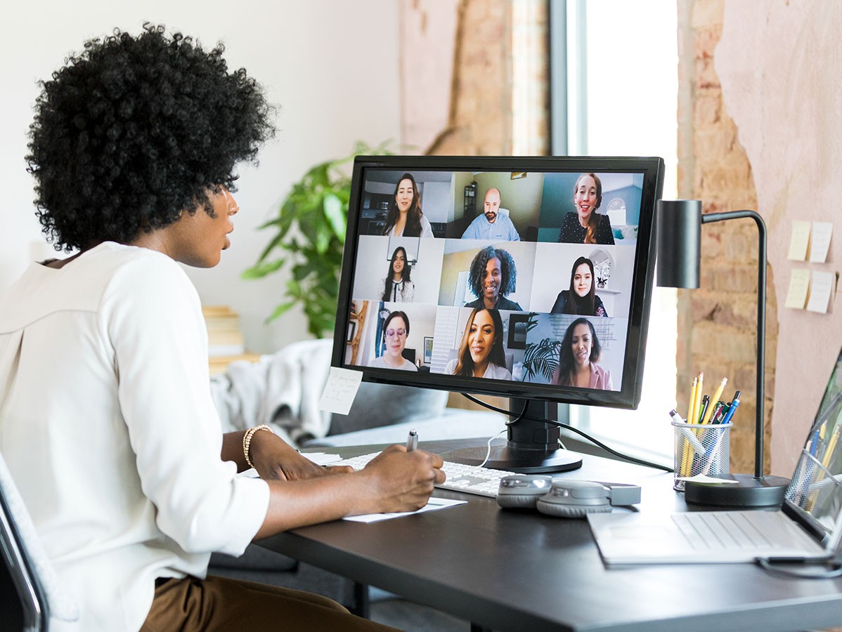 Woman sitting at a desk looking at her computer screen. She is in a virtual meeting and 9 people appear on the screen.