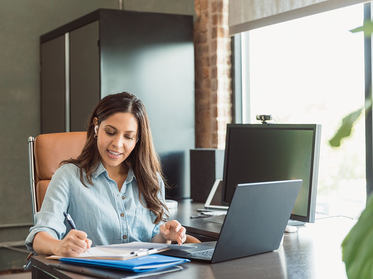 Woman in an office writing notes and smiling. A laptop and computer monitor are on the desk.