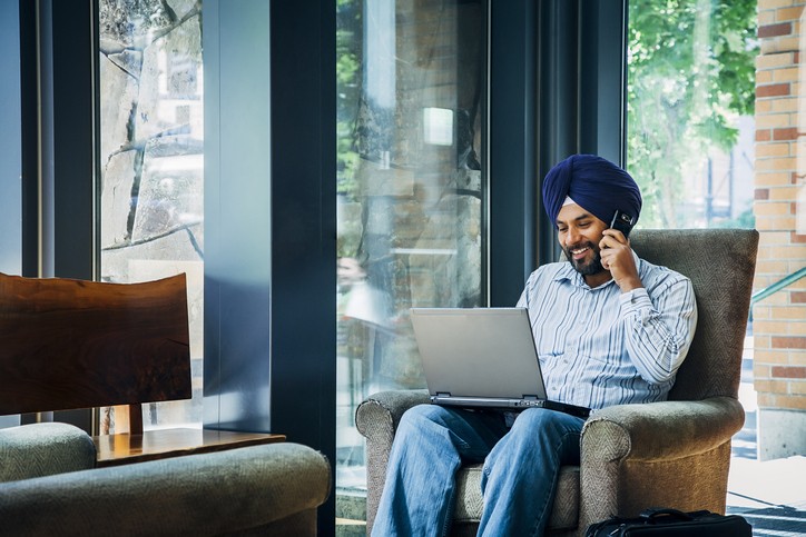 A smiling South Asian man using a laptop at home, while talking on a cell phone