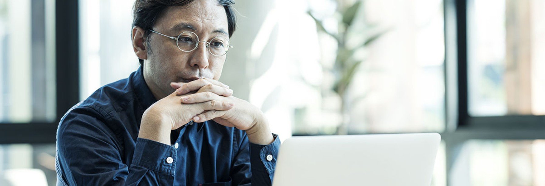 Man with glasses in a blue shirt sitting at a table with coffee looking at his laptop.